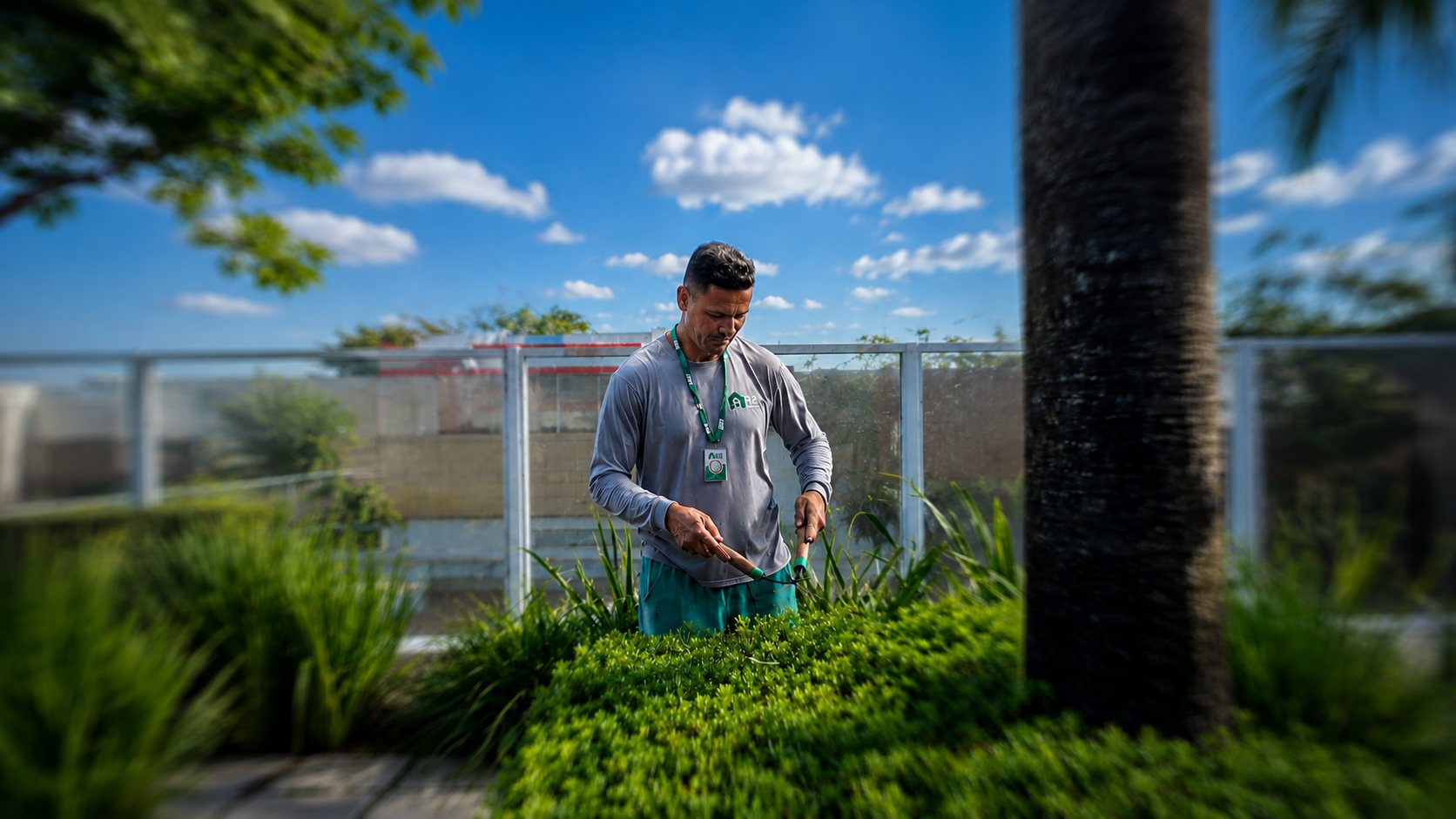 Profissional uniformizado da R2 Service aparando arbustos em jardim corporativo elevado, sob céu azul